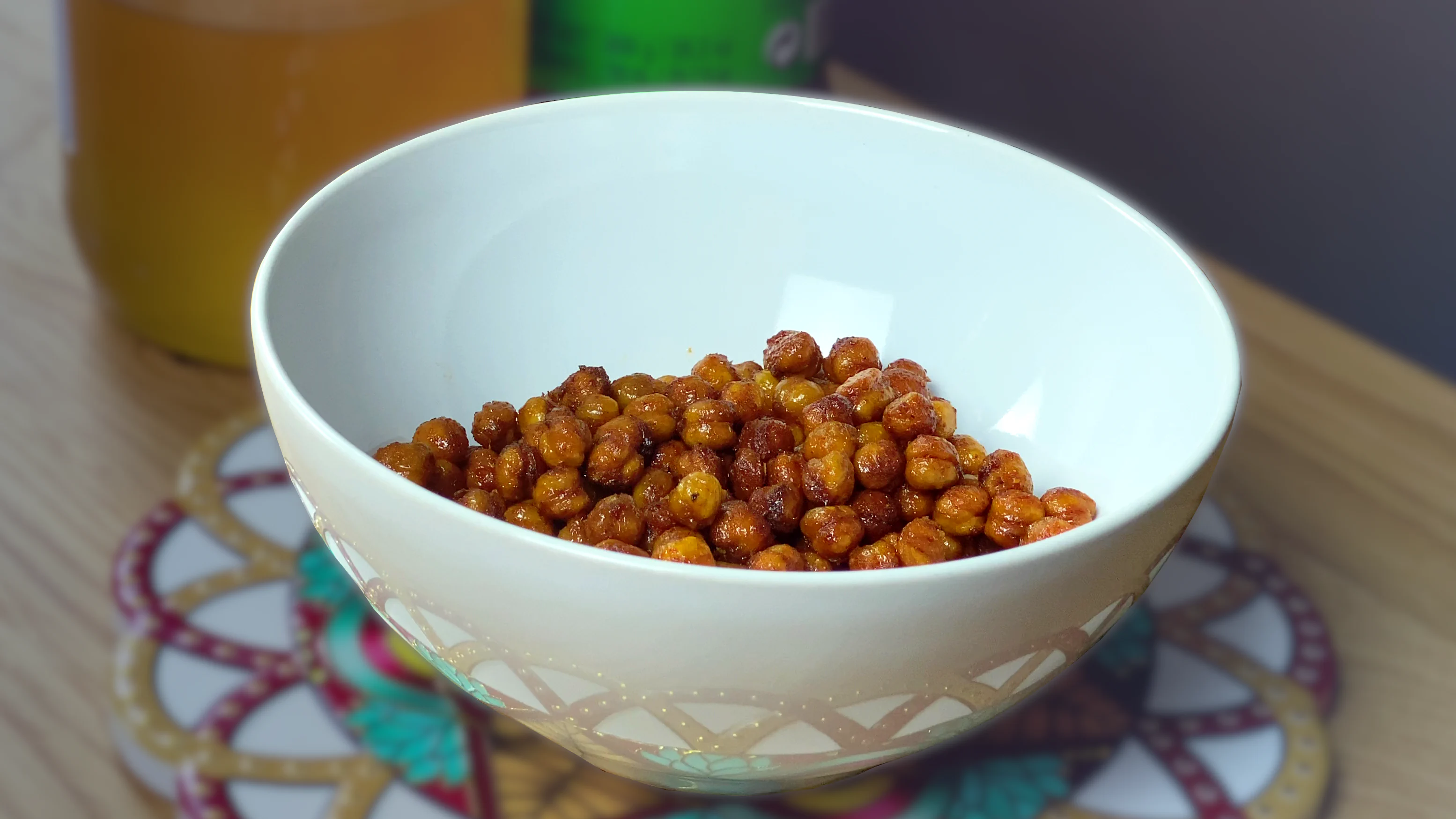 A white bowl filled with golden-brown roasted chickpeas, placed on a colorful patterned coaster on a wooden surface. In the background, there is a blurred honey jar and sriracha sauce bottle.