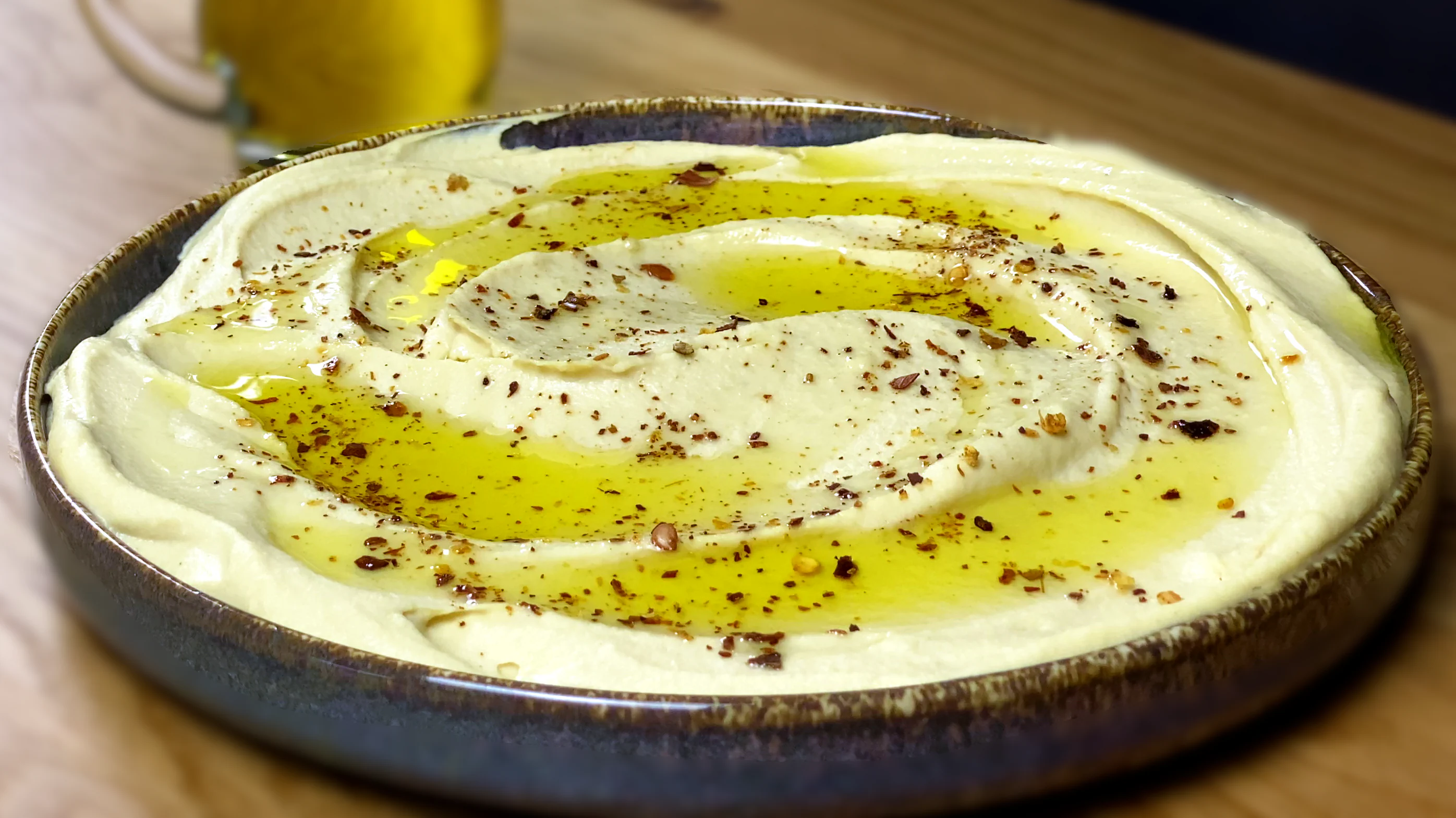 Creamy hummus topped with olive oil and chili flakes on a blue ceramic plate, with a blurred olive oil bottle in the background.