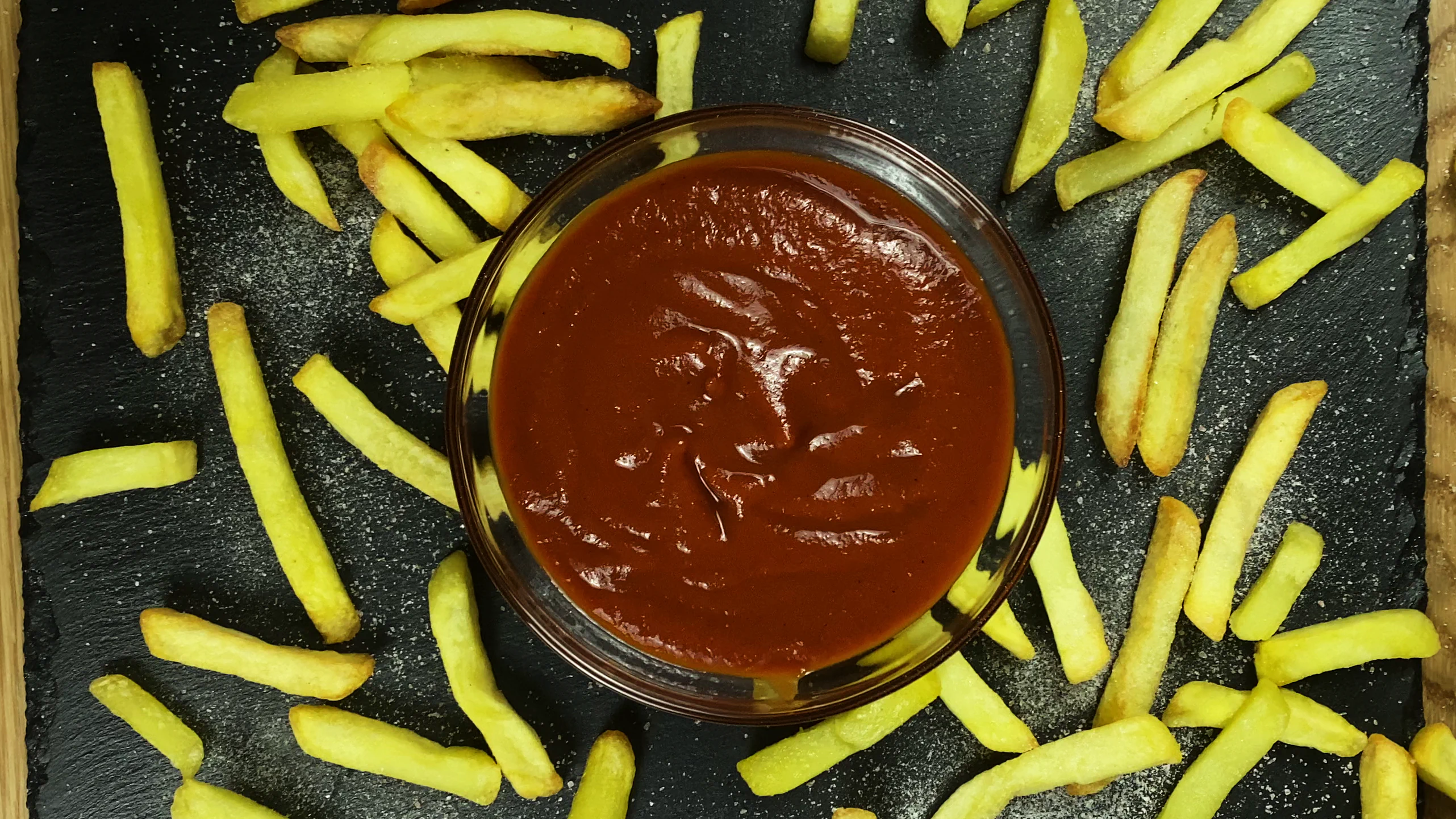 Top-down view of a glass bowl filled with ketchup, surrounded by golden French fries scattered on a dark slate surface.