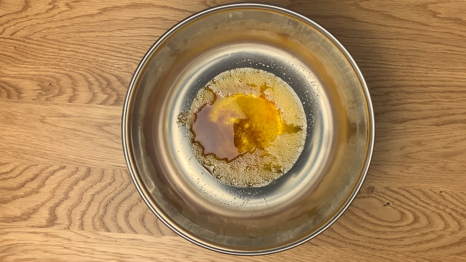 op-down view of a shallow stainless steel bowl on a wooden surface, containing melted brown butter with golden and amber tones.