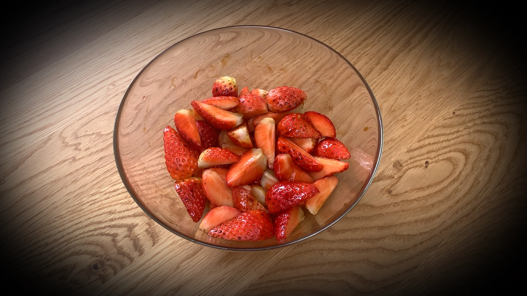 Bowl of freshly cut strawberries in a clear glass mixing bowl on a wooden surface, with the fruit appearing juicy and ready for preparation in a strawberry-ricotta dessert.