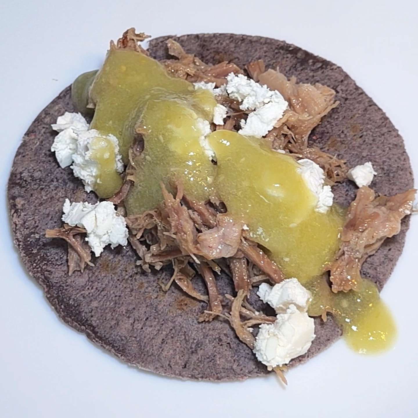 Assembled carnitas taco with pulled pork, white cheese, and salsa verde on a dark blue corn tortilla.