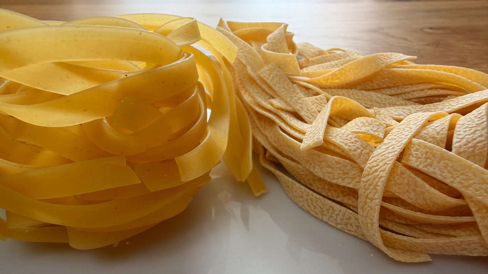 Close-up of two uncooked tagliatelle pasta on a white plate - on the left smooth and glossy, on the right slightly rough and textured - both in warm yellow tones under soft natural light.