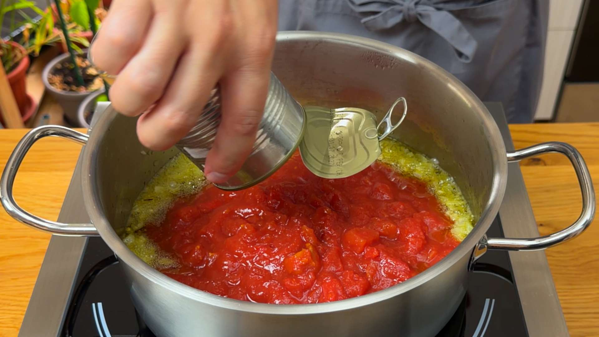 A person pouring canned peeled tomatoes into a stainless steel pot with sautéed garlic and oil.