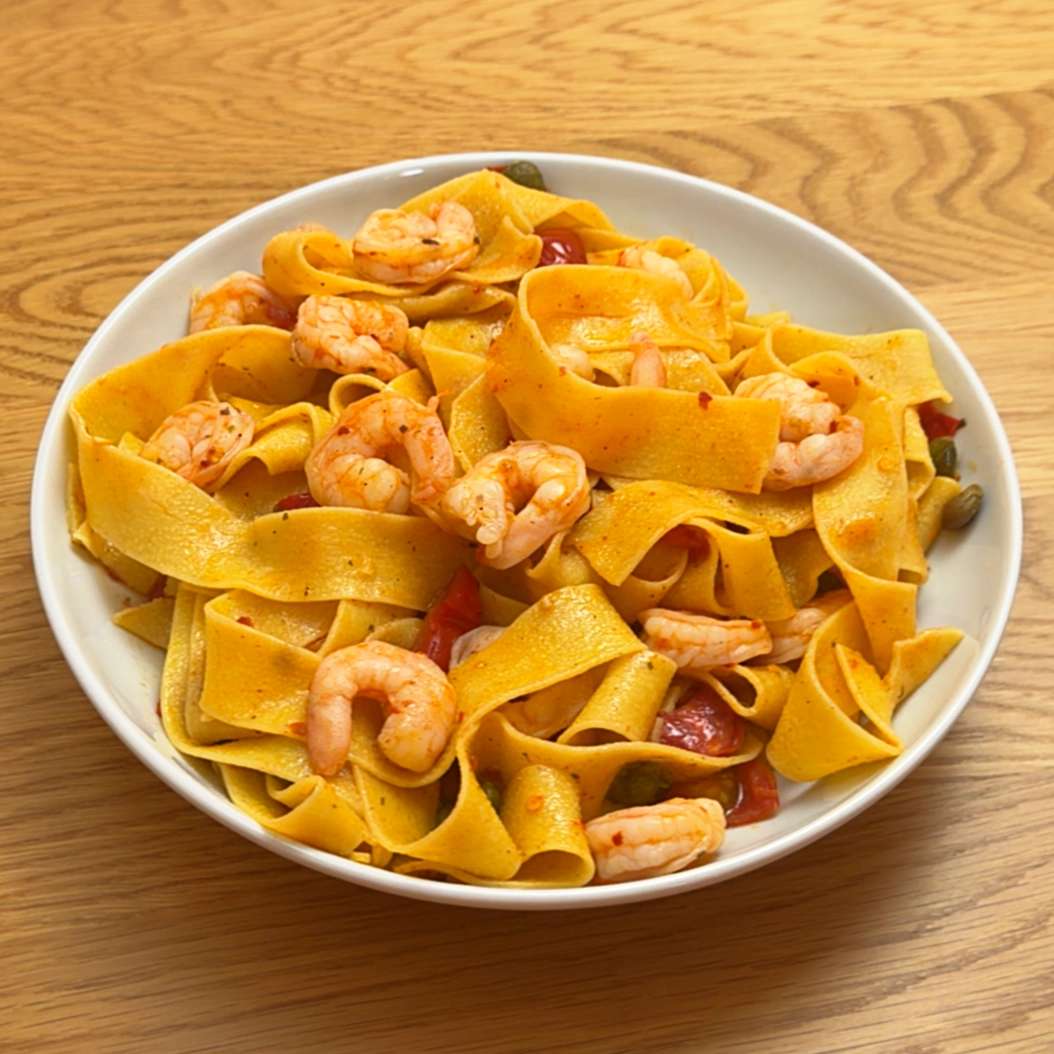 A bowl of wide pappardelle pasta with shrimp, capers, and cherry tomatoes on a wooden table.