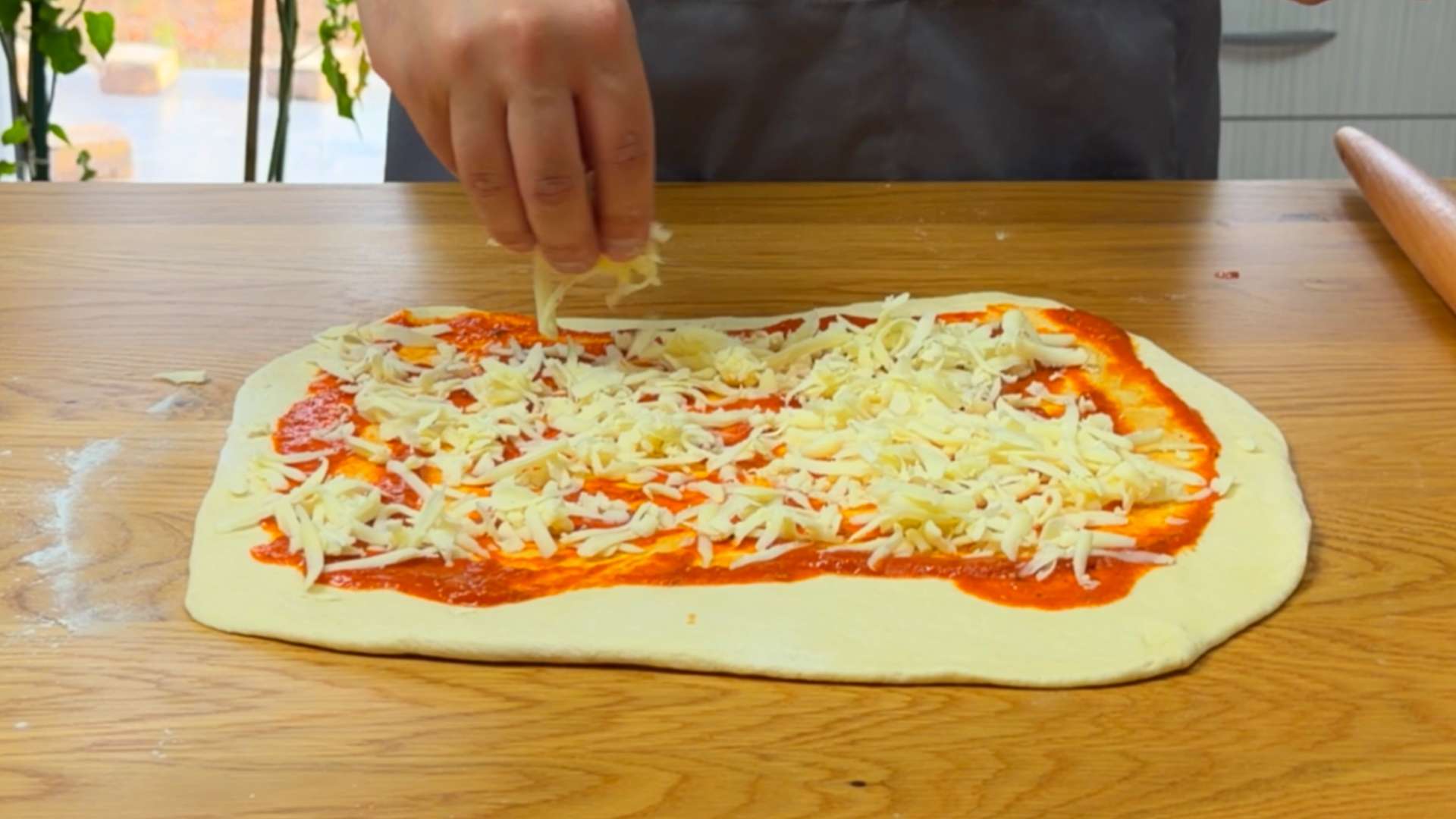 A person sprinkling shredded mozzarella cheese over tomato sauce on rolled-out pizza dough.