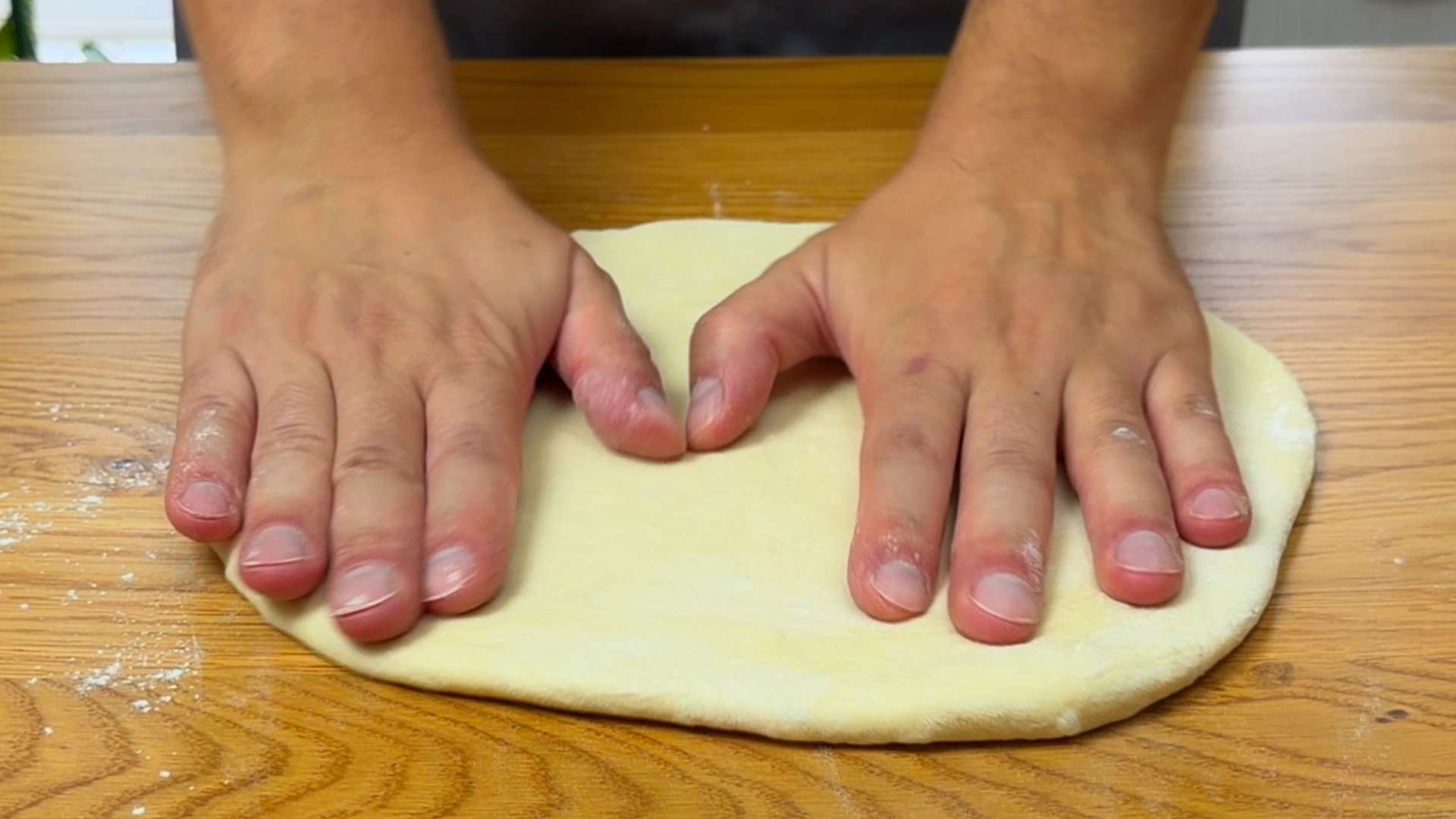 Two hands pressing and flattening homemade pizza dough on a wooden countertop.