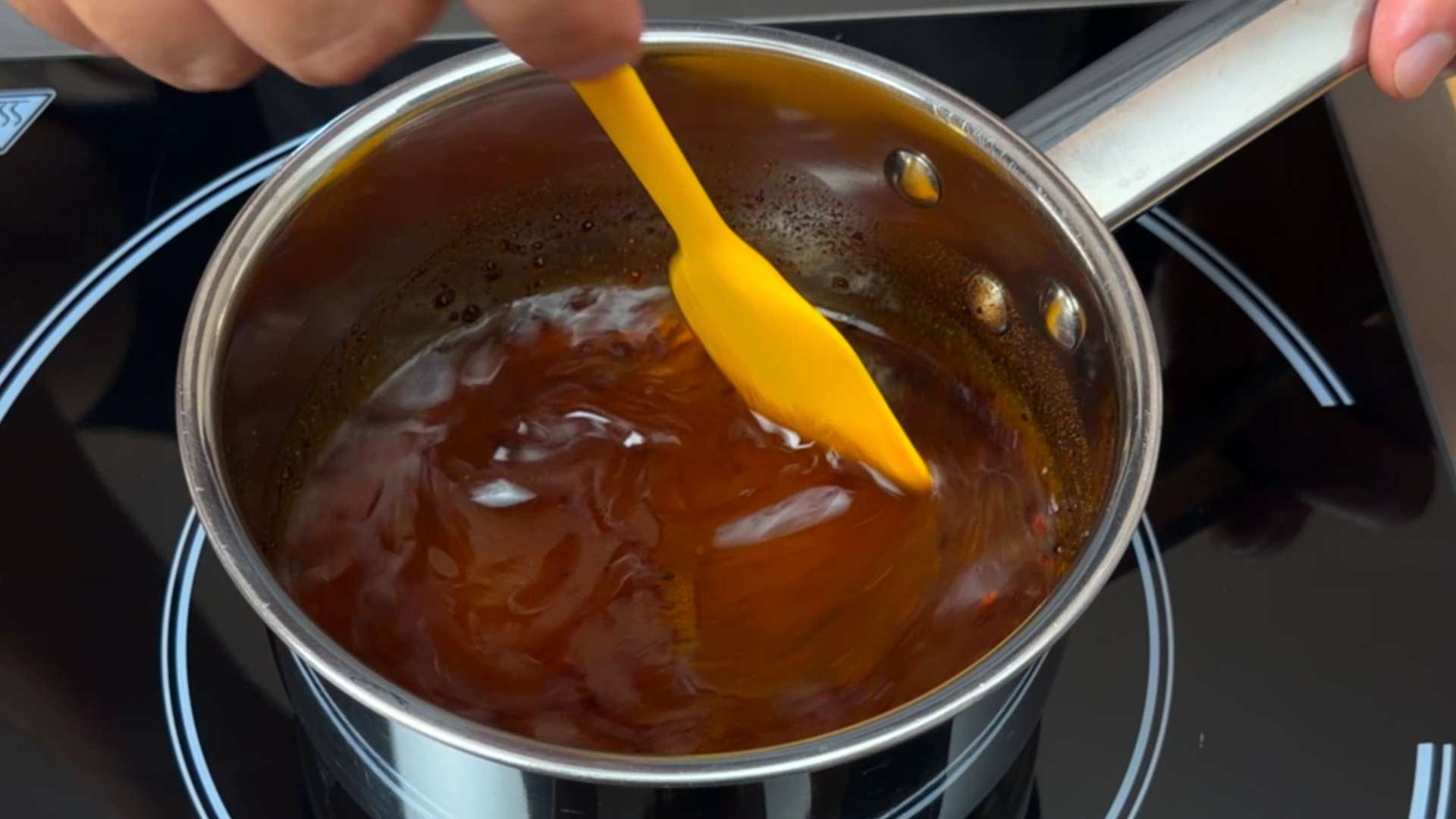 Stirring amber hot honey with a yellow silicone spatula in a small stainless steel saucepan on an induction stove.