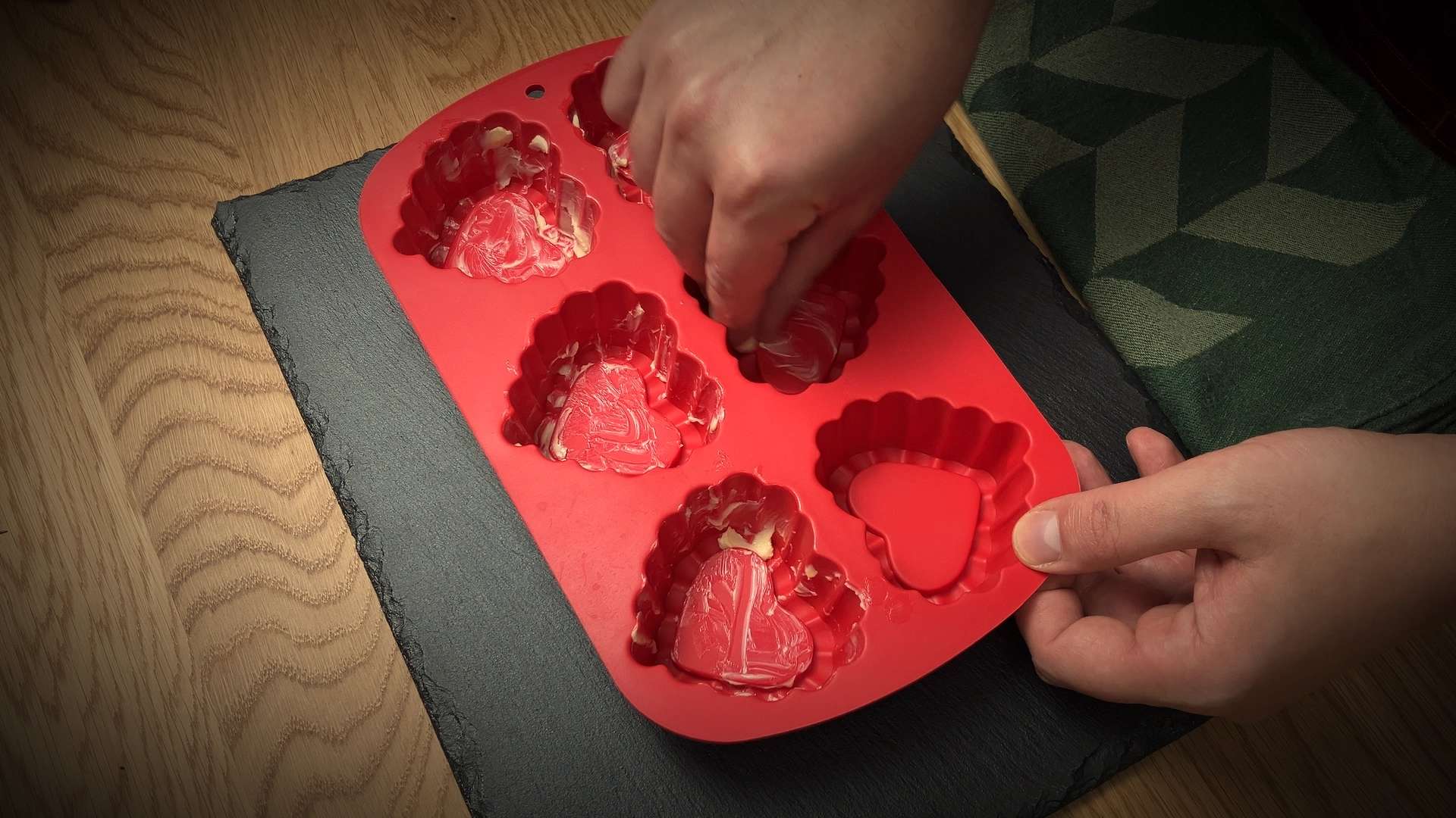 Hands spreading a batter inside a red silicone heart-shaped baking mold on a wooden surface, preparing molds for baking.