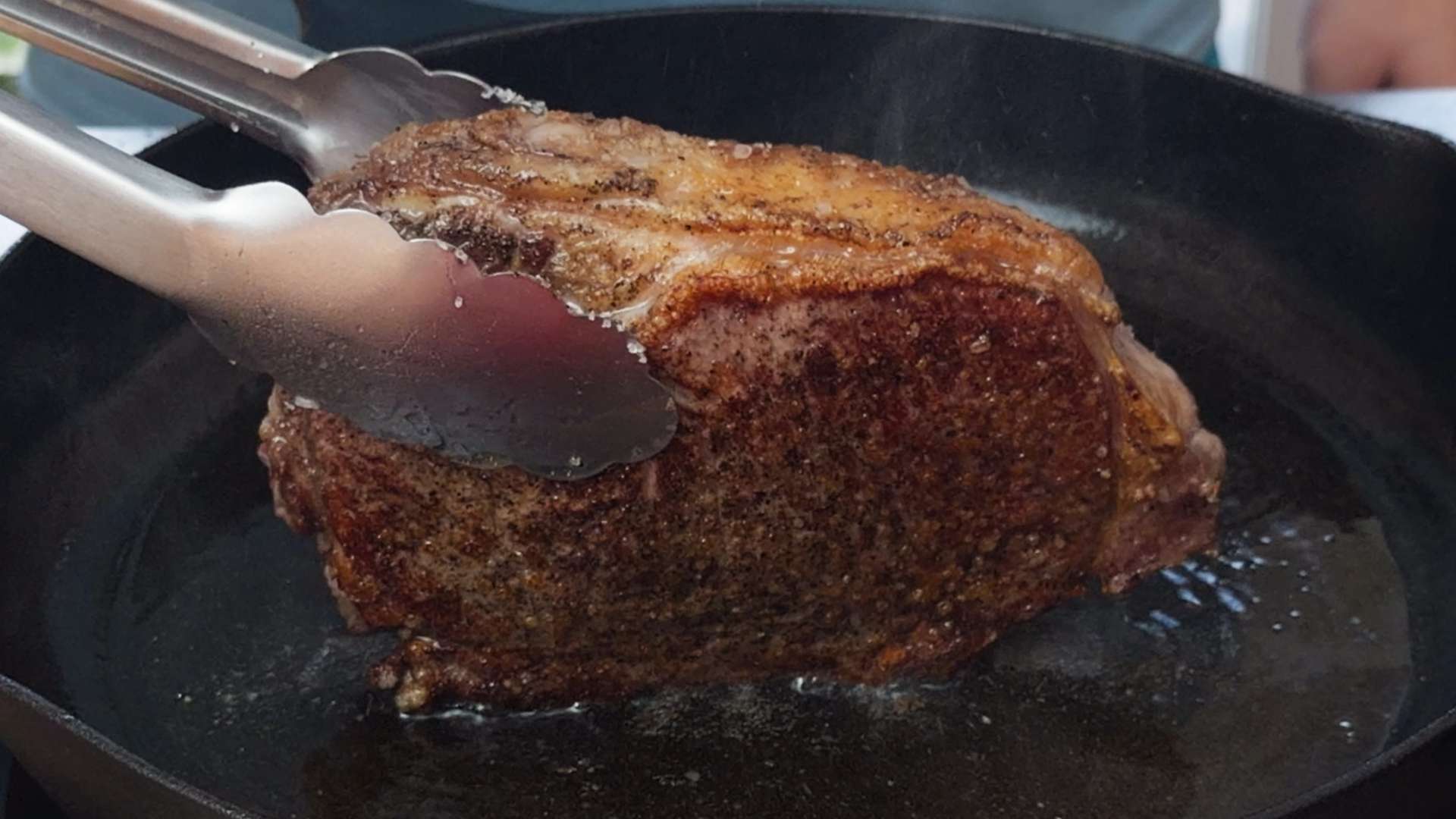 Close-up of a thick ribeye steak searing in a cast iron skillet, held with stainless steel tongs. The steak has a deep golden-brown crust with rendered fat along the edge, showcasing the perfect sear before finishing the cook.