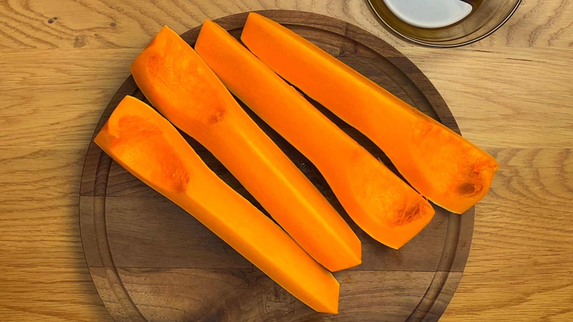 Four long, peeled, and sliced raw butternut squash quarters resting on a round wooden cutting board.