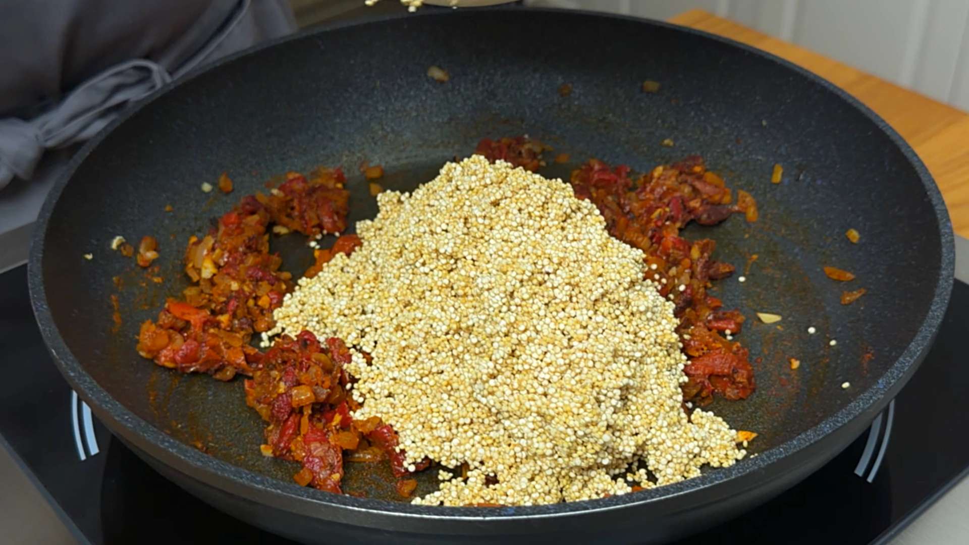 Uncooked grain quinoa added on top of sautéed onions and harissa paste in a large black pan.