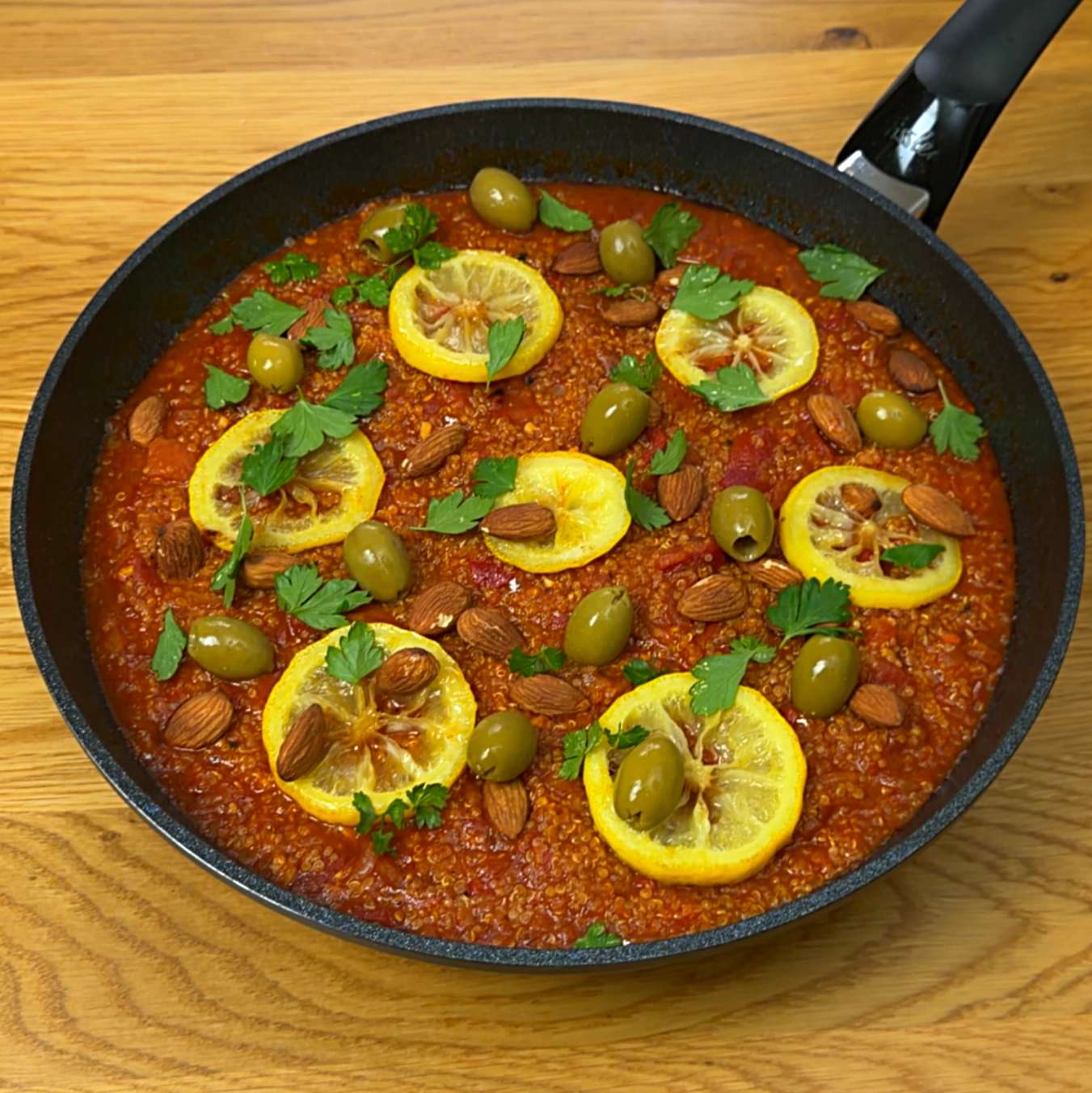 Top-down view of cooked harissa quinoa dish garnished with lemon slices, whole almonds, green olives, and fresh parsley in a skillet.