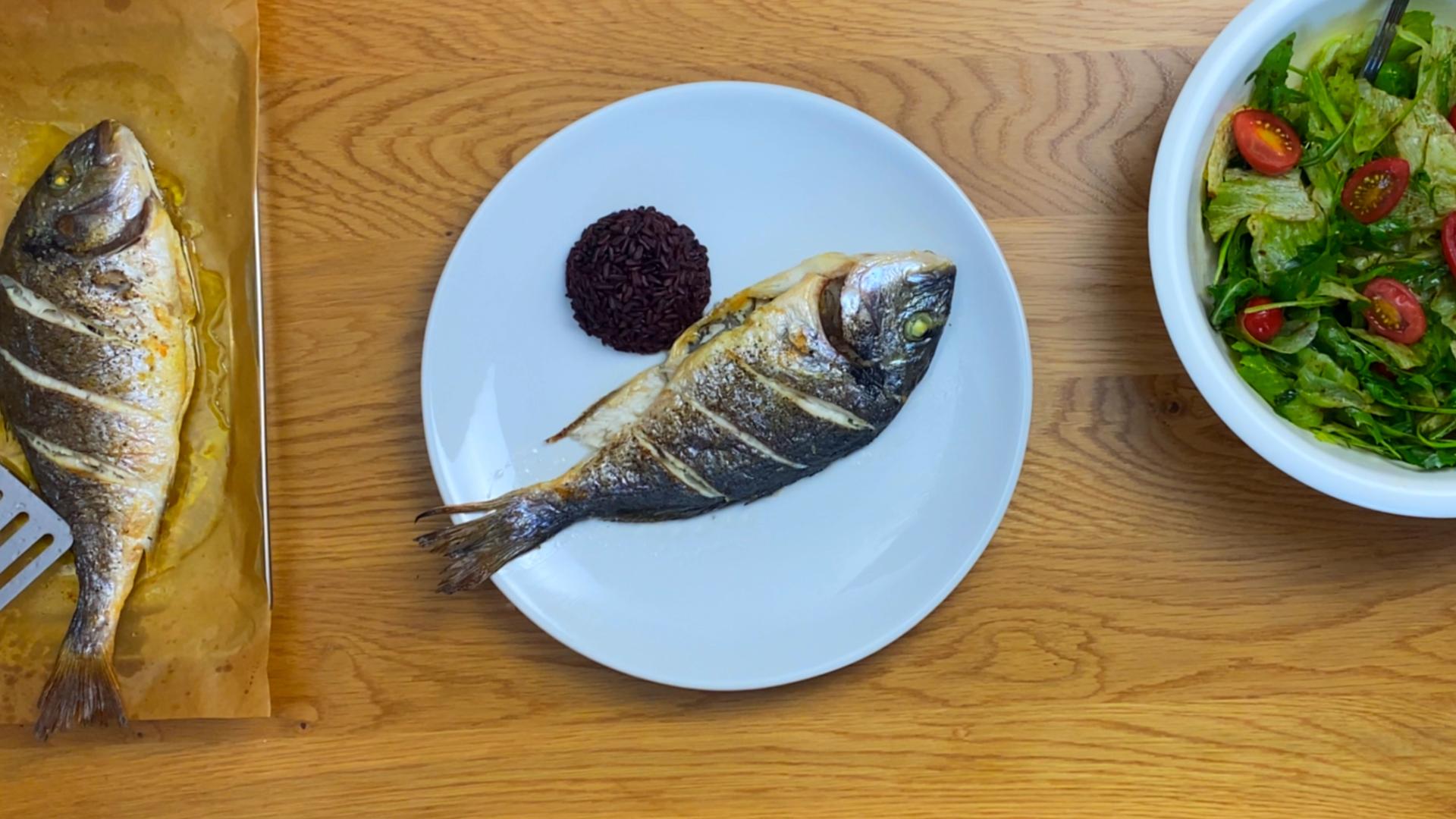 Whole oven baked sea bream served with black rice and fresh green salad on a wooden table.