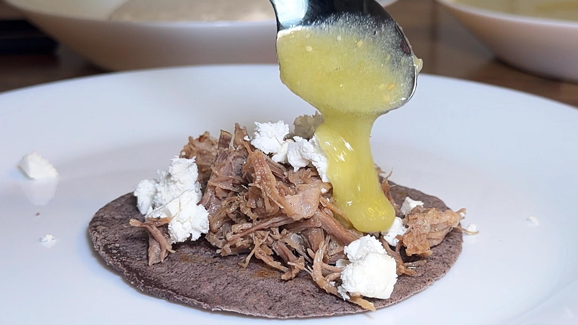 Spoon pouring green salsa verde onto a carnitas taco with crumbled cheese on a blue corn tortilla.