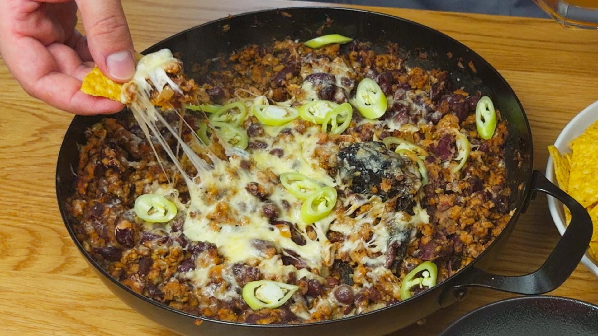 One-pan cheesy beef and kidney bean nacho dip topped with sliced green chili peppers, served in a carbon steel pan with tortilla chips.
