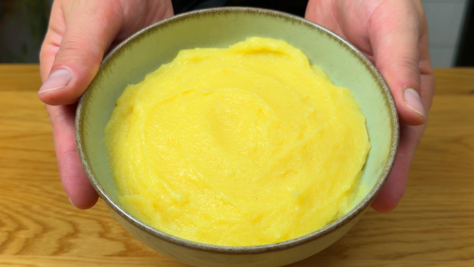 A ceramic bowl filled with smooth cooked corn flour polenta, held by two hands above a wooden surface.