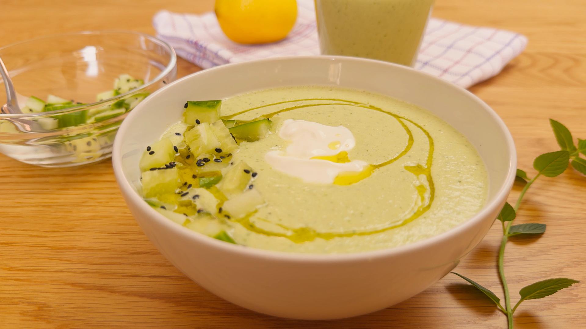 Bowl of chilled cucumber gazpacho garnished with diced cucumber, yogurt, olive oil, and black sesame seeds on a wooden table.