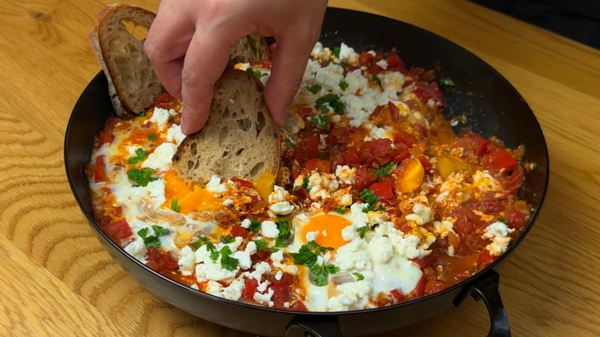 A hand dipping crusty bread into a skillet of shakshuka topped with feta and herbs.