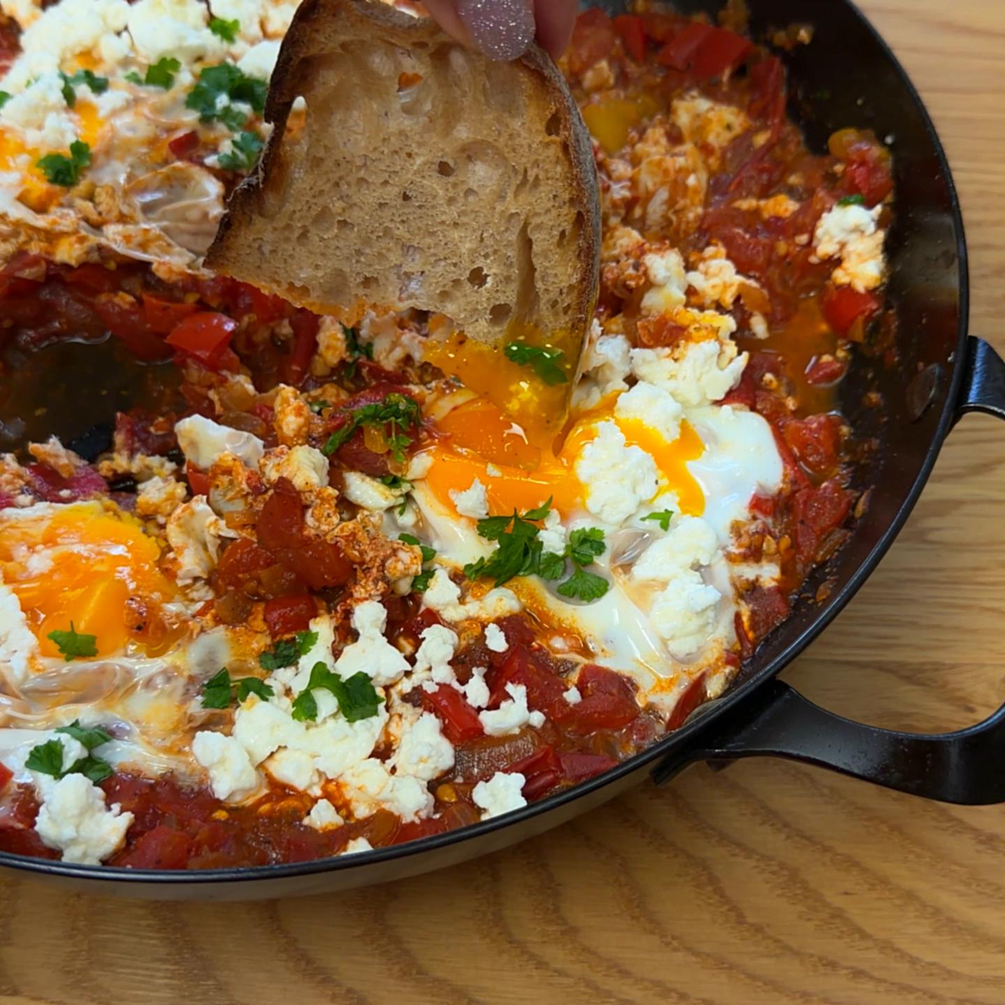 A close-up of bread breaking a runny egg yolk in a skillet of shakshuka.