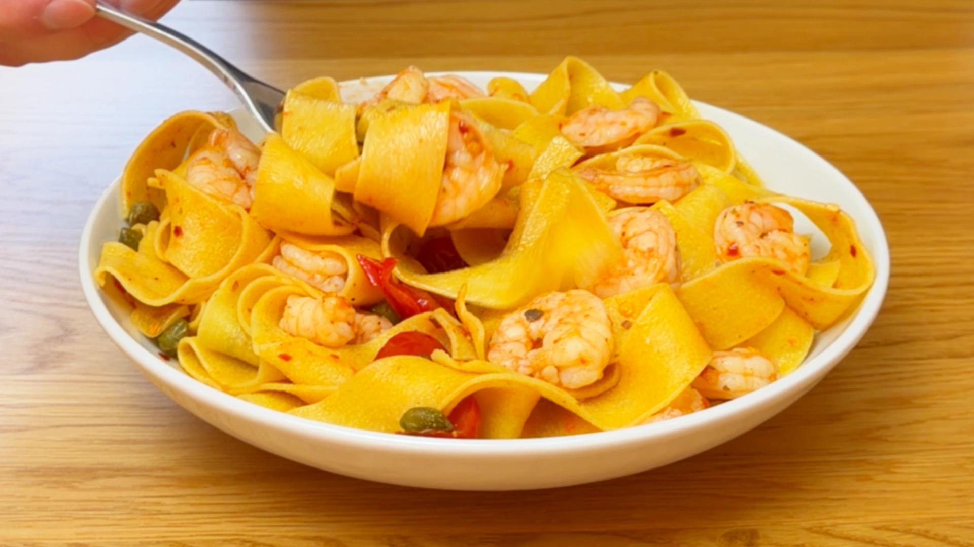 A fork lifting wide pappardelle pasta with shrimp from a white bowl.