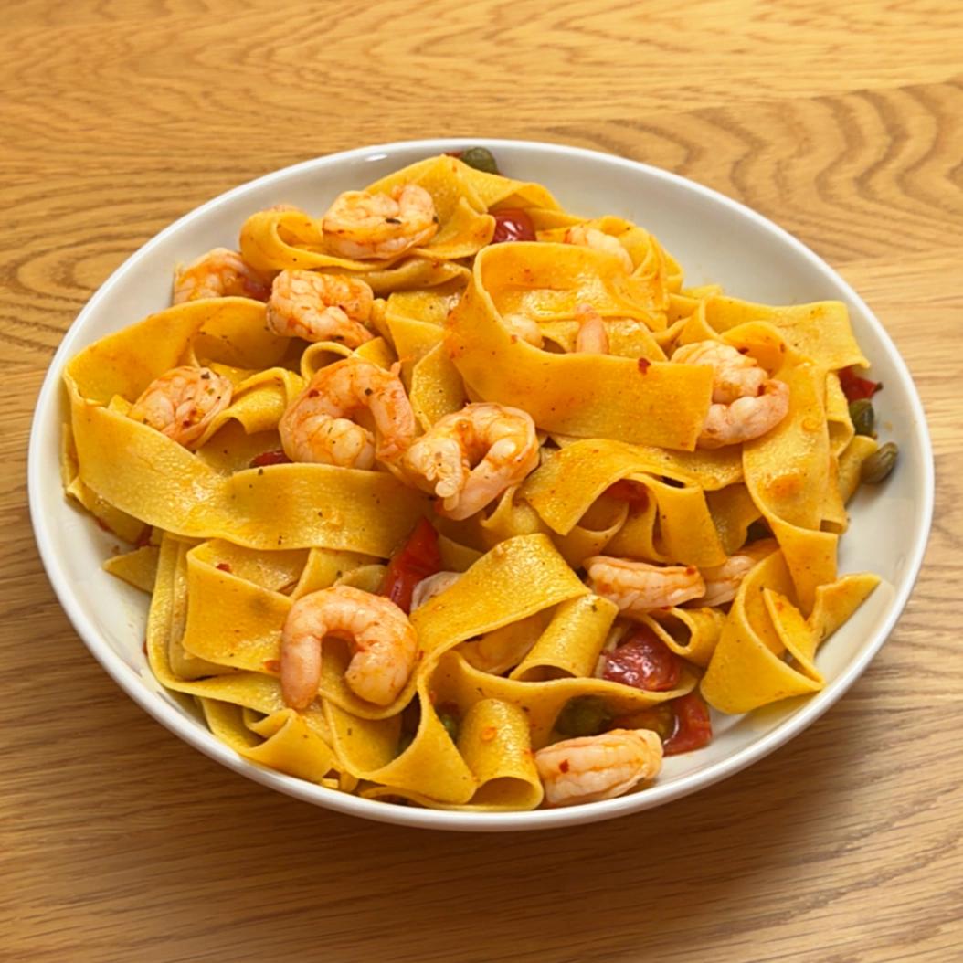 A bowl of wide pappardelle pasta with shrimp, capers, and cherry tomatoes on a wooden table.