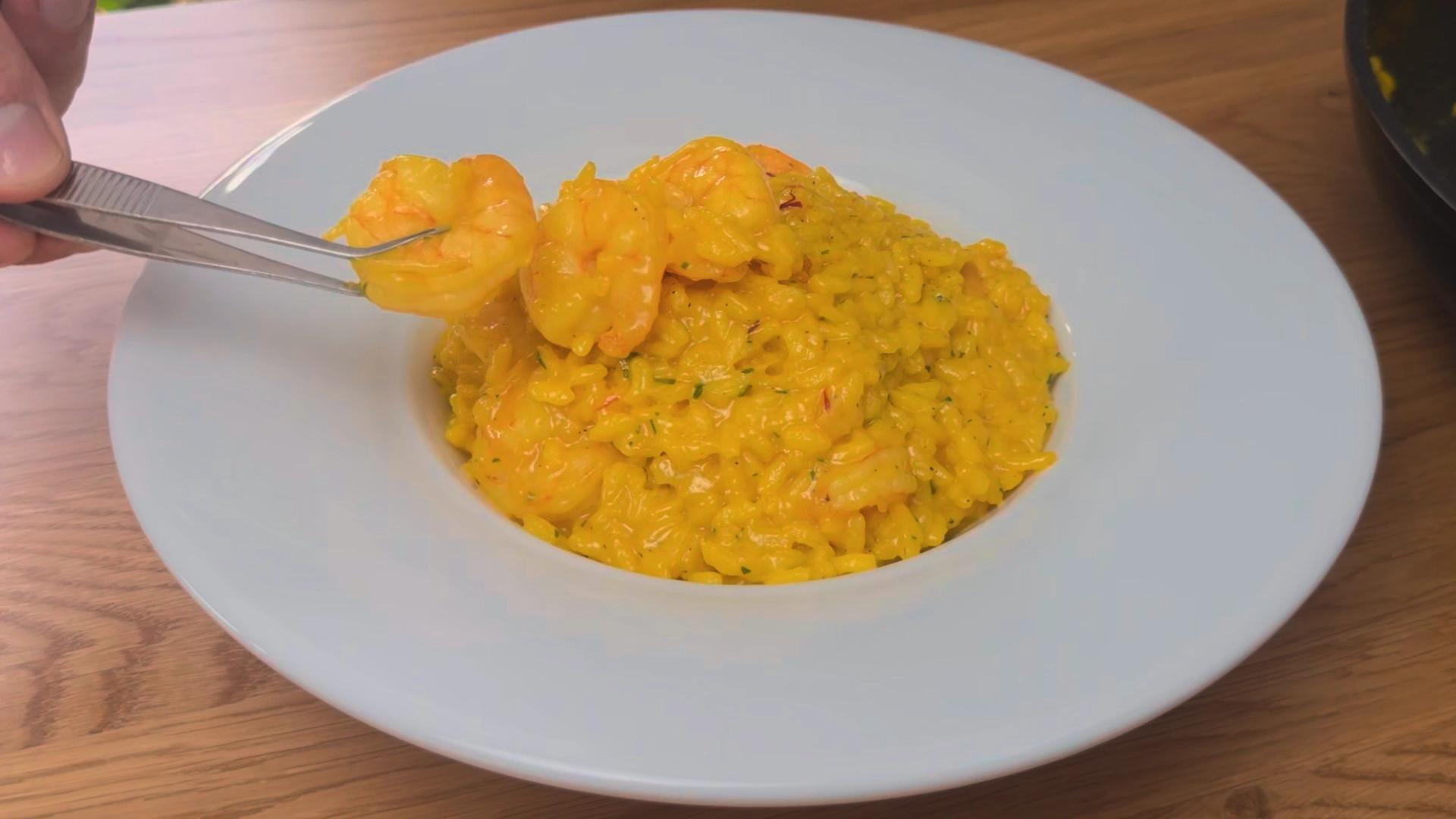 Creamy saffron shrimp risotto served in a white bowl with tender shrimp and a rich golden texture, while tweezers lift a shrimp above the dish.