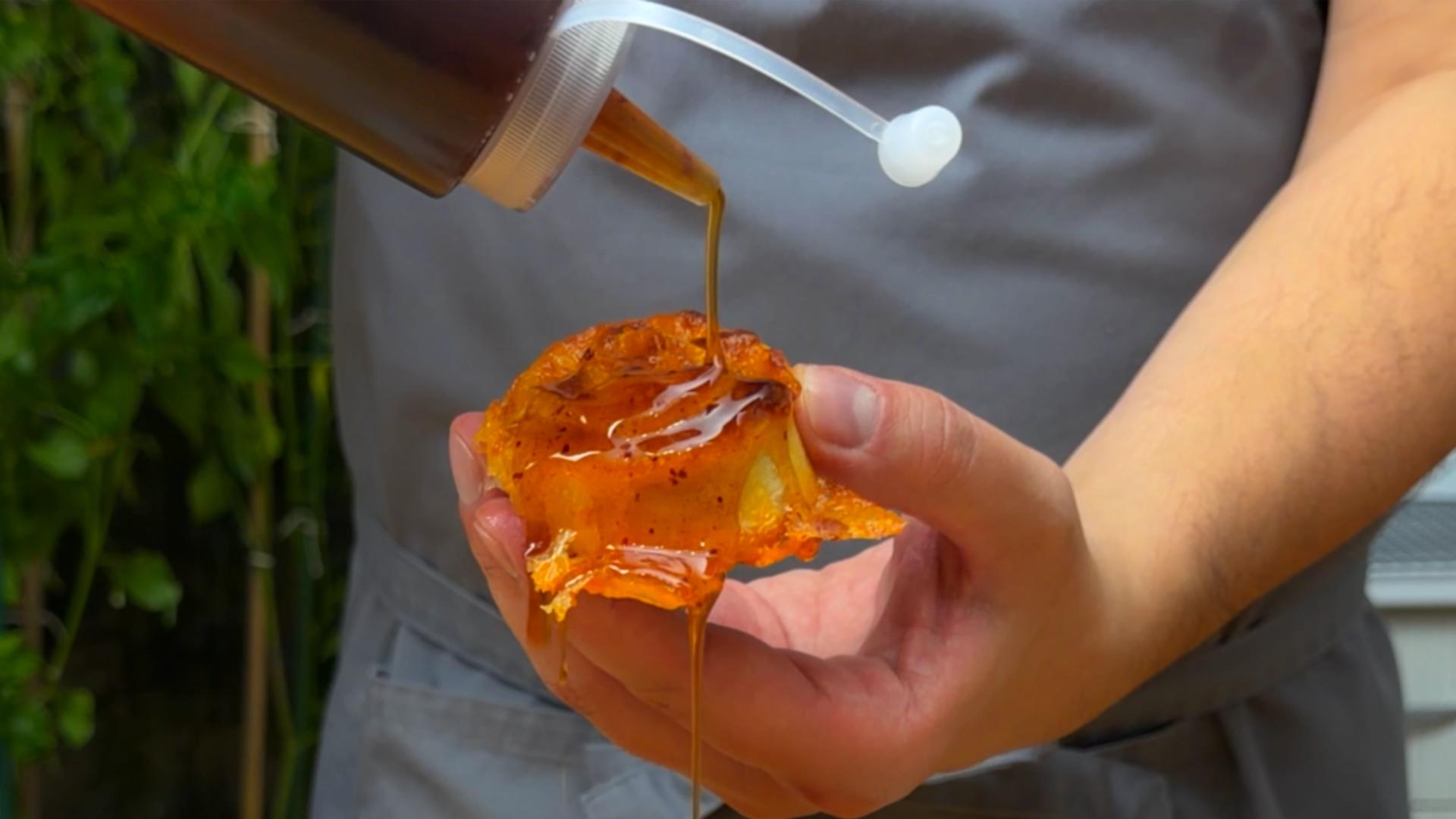 Drizzling golden-brown hot honey from a plastic squeeze bottle onto a crispy pastry held in a hand.