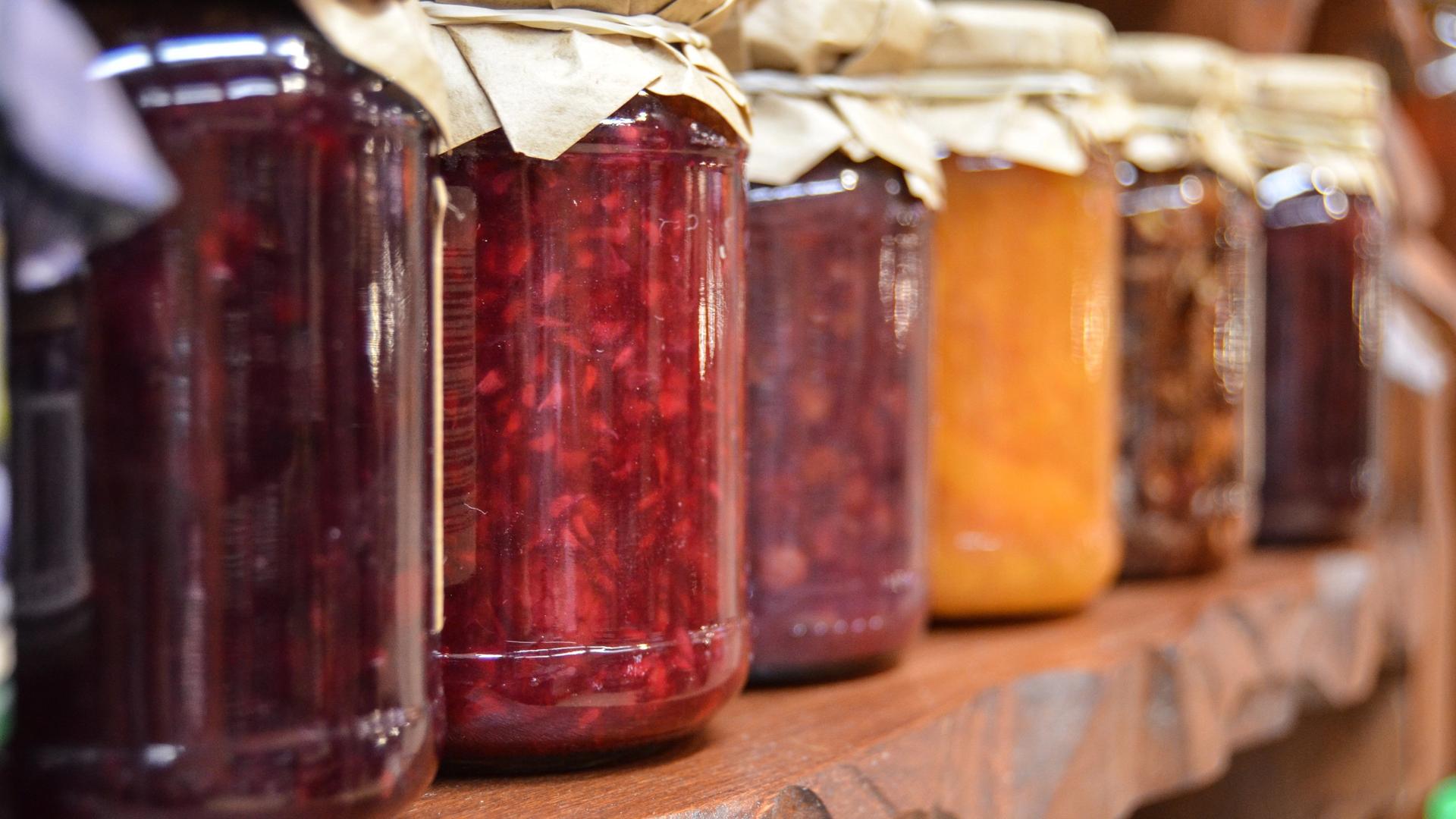 Jars of homemade preserves in various colors, including red, purple, orange, and dark berry, arranged on a wooden shelf.