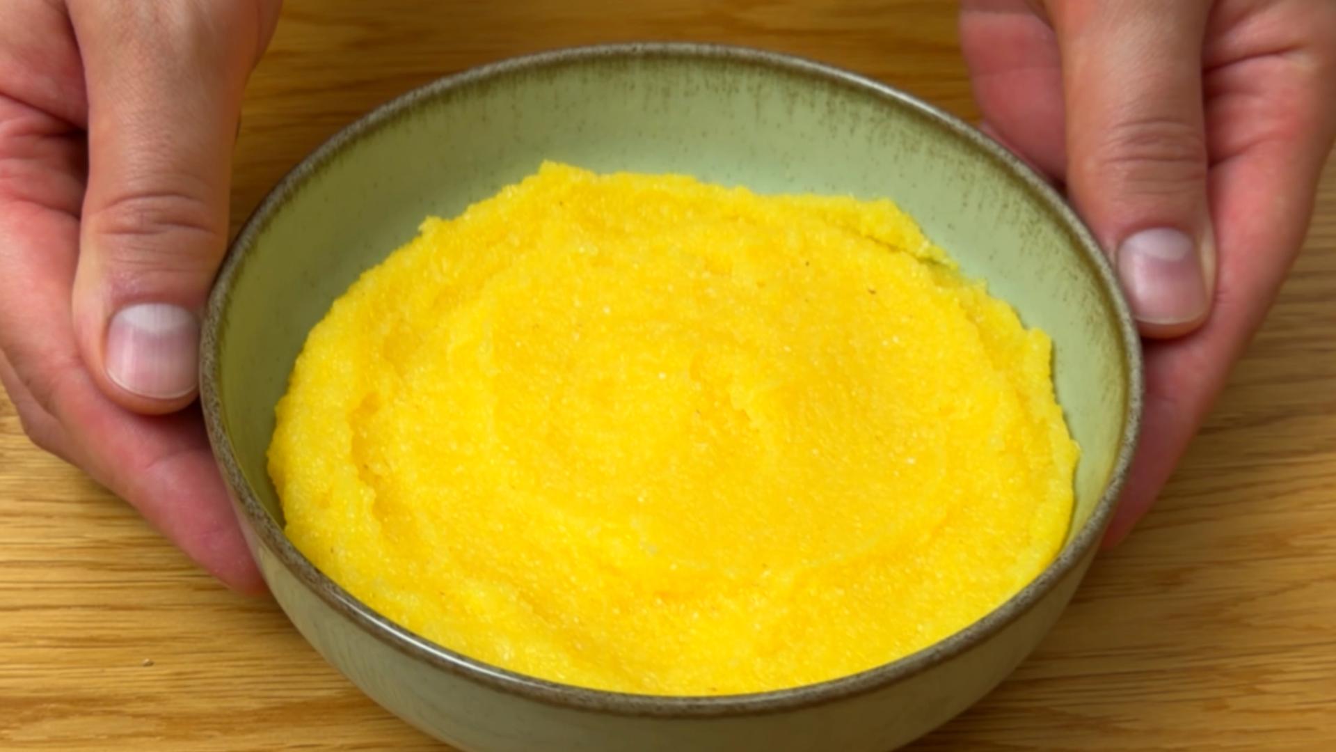 A ceramic bowl filled with smooth cooked instant polenta, held by two hands on a wooden surface.