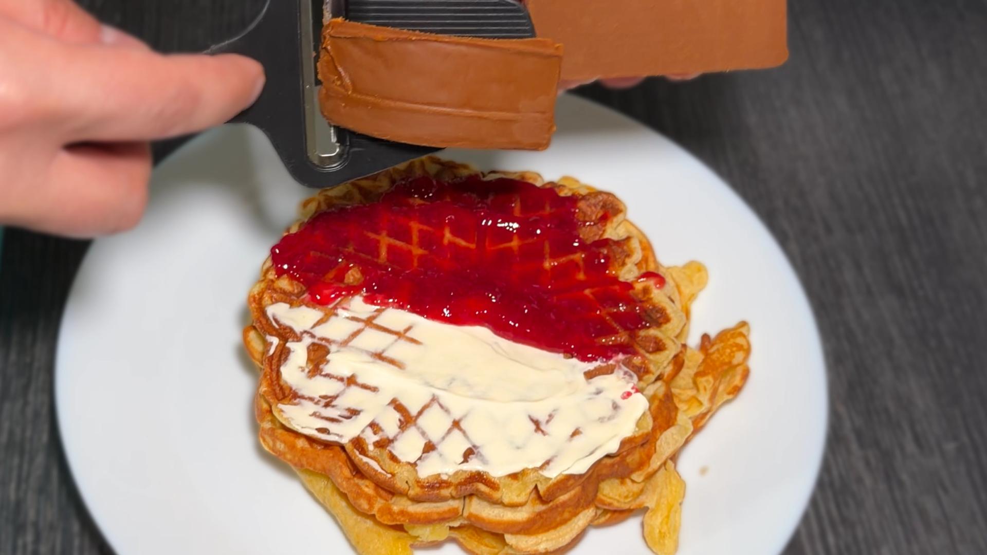 Top view of stacked waffles topped with red jam and sour cream, with thin slices of Norwegian brown cheese (brunost) being shaved over the top.