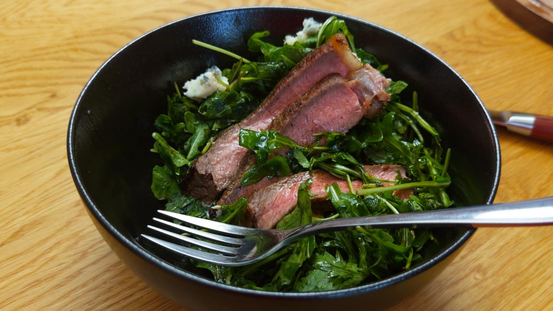 Medium-rare sliced steak served over a fresh arugula salad with crumbled Gorgonzola cheese, lightly dressed and presented in a black bowl on a wooden table. A fork rests in the bowl, highlighting the juicy steak and vibrant greens.