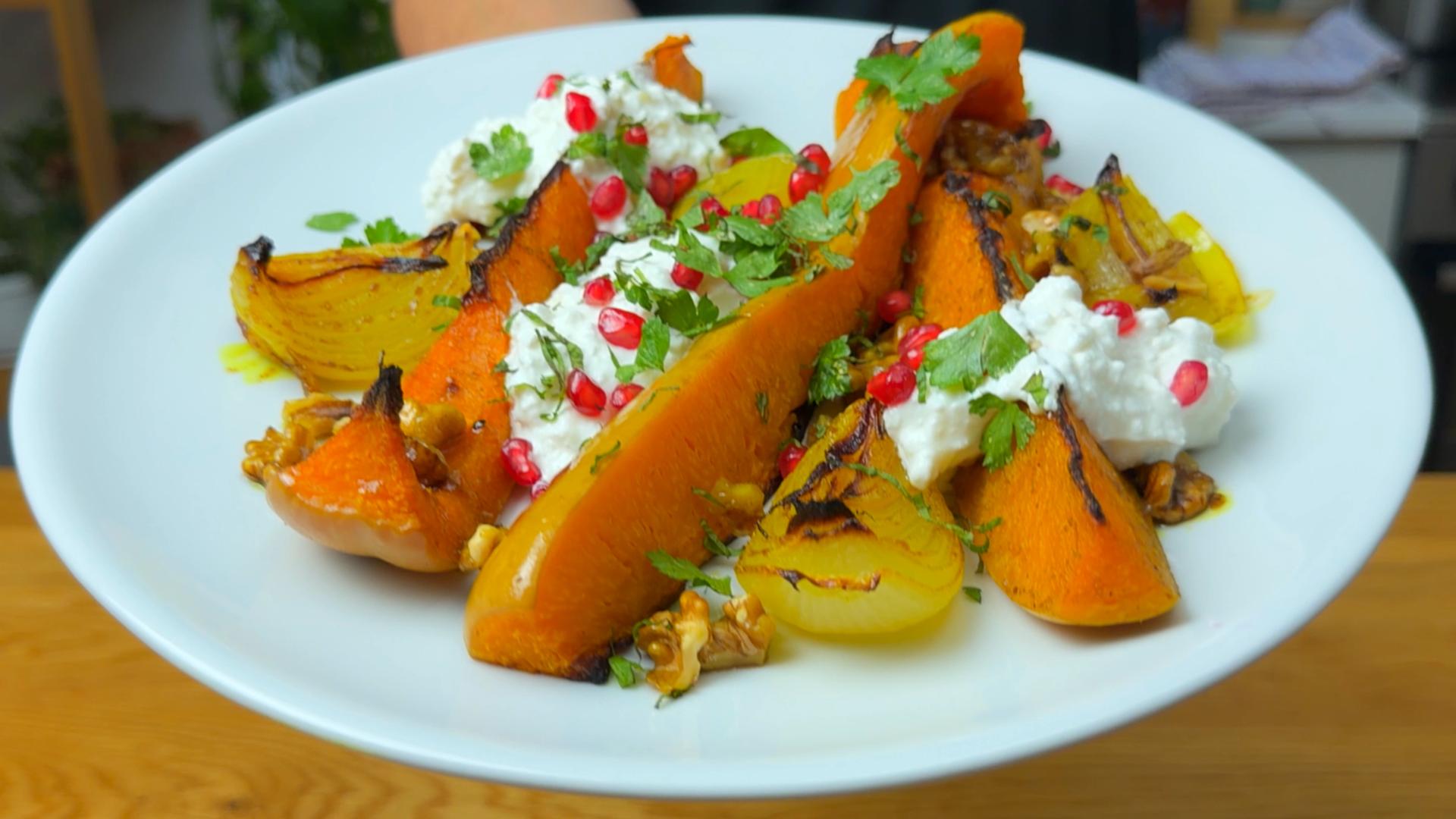 Roasted butternut squash wedges served with whipped feta yogurt sauce, pomegranate seeds, walnuts, and fresh herbs on a white plate.