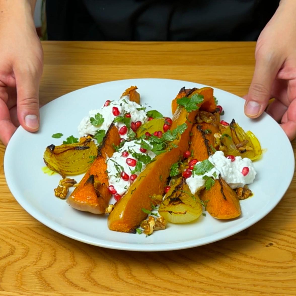 Hands holding a white plate topped with roasted butternut squash wedges, whipped feta sauce, pomegranate seeds, and walnuts.