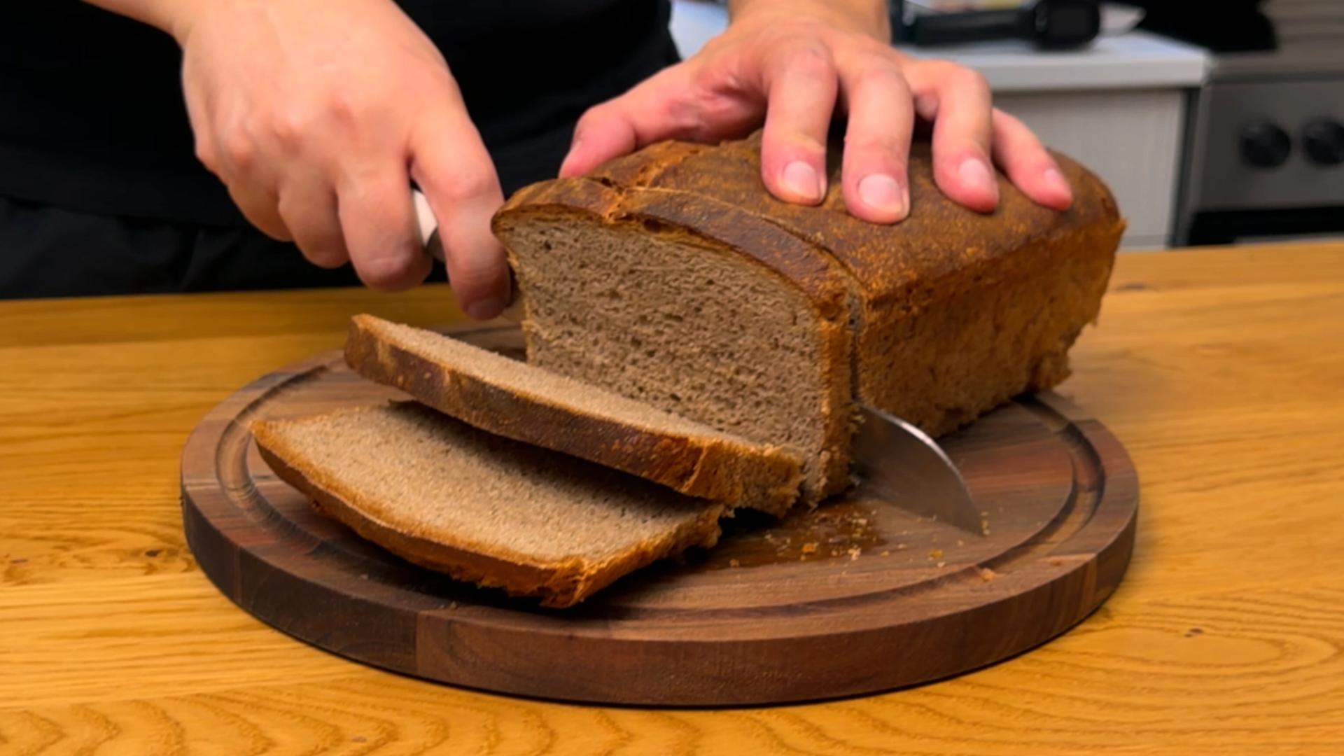 A close-up shot of hands using a knife to slice a loaf of whole wheat bread on a cutting board, with visible bread crumbs and grain texture.